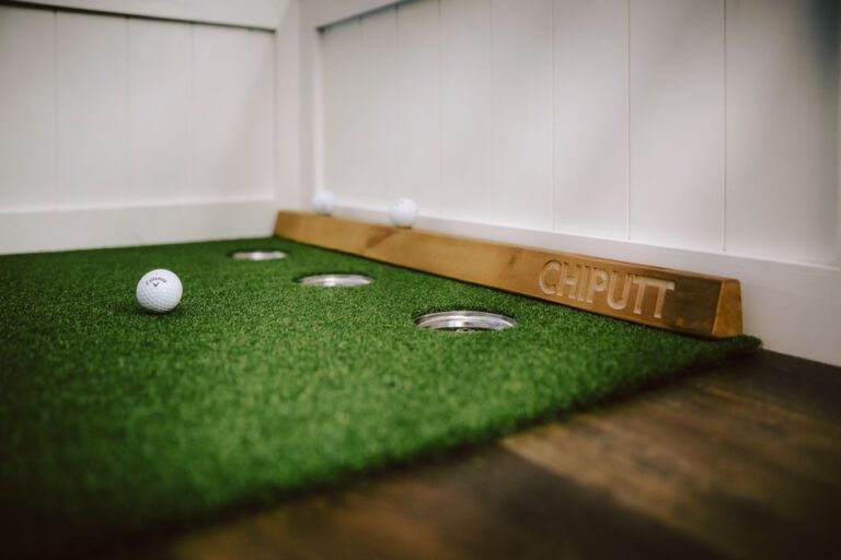 Two people practicing putting on a green mat indoors with household objects as targets.