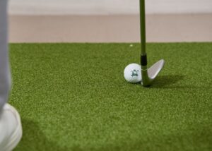 A golfer swings an iron on moist spring grass, with a golf ball and divot flying against a green fairway backdrop.