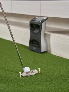 A group of people swinging golf clubs in an indoor simulator bay, watching their virtual shots on a large screen.