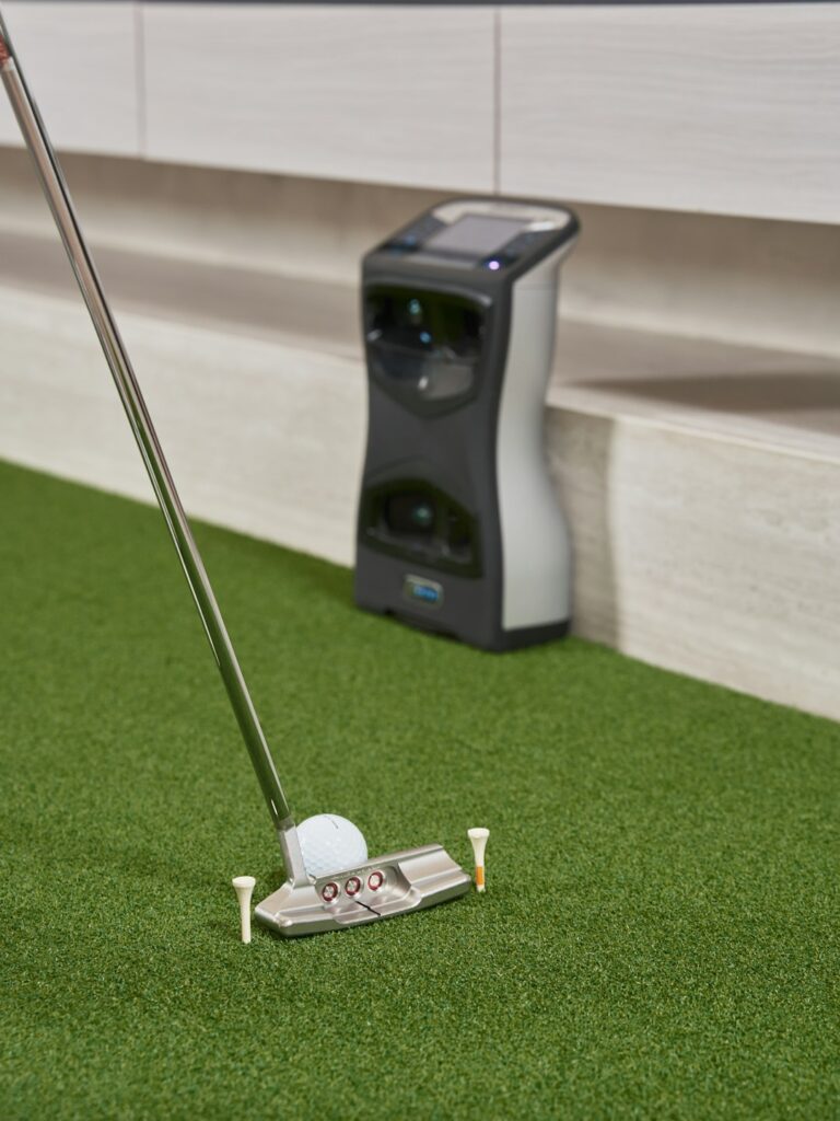 A group of people swinging golf clubs in an indoor simulator bay, watching their virtual shots on a large screen.